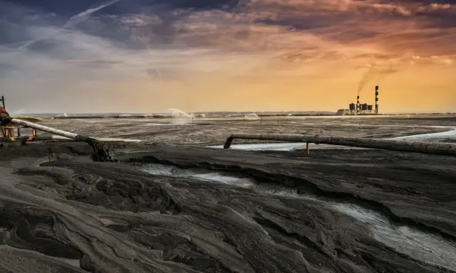 Krajobraz postindustrialny, gdzie rury odprowadzają odpady, a w oddali widać elektrownię. Woda i ziemia tworzą surowy obraz, symbolizujący proces rekultywacji.
