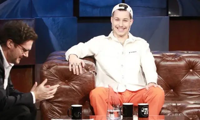 Zabson, z uśmiechem na twarzy, siedzi na kanapie w studiu. Obok niego siedzi mężczyzna w okularach.
