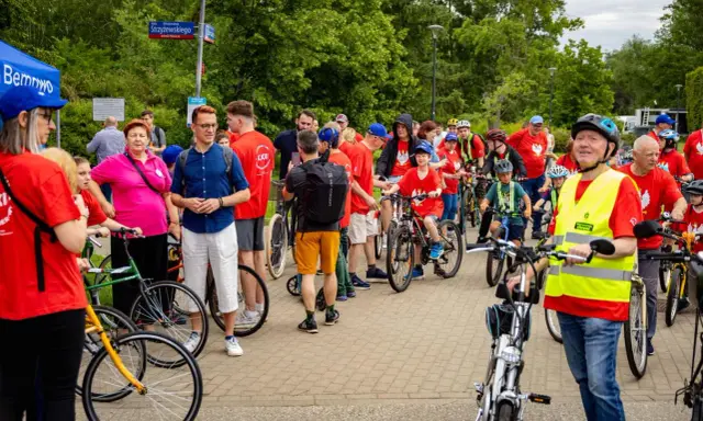 Uczestnicy rajdu rowerowego w czerwonych koszulkach, dzieci i dorośli, gotowi do startu.