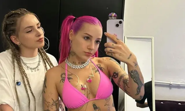 Dwie młode kobiety, jedna z różowymi włosami i tatuażami, druga z warkoczami, pozują do selfie.