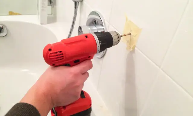 Une main tient une perceuse rouge pour percer du carrelage blanc, avec du ruban adhésif pour guider le foret.