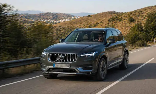 Volvo XC90 oscuro, uno de los suv mas fiables, circulando por una carretera rural con colinas verdes de fondo.