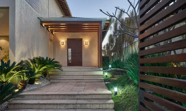 Un portico moderno con porta in legno scuro, illuminazione soffusa e piante rigogliose. Un esempio di come abbellire un portico con stile e natura.