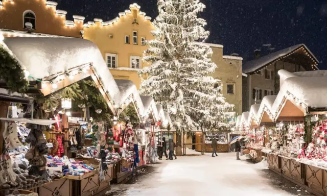 Mercatini di Natale nel Nord Italia, con bancarelle innevate, un grande albero illuminato e case decorate. Magia invernale.