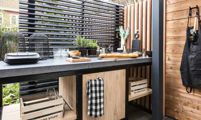 Cucina all'aperto fai da te con piano in cemento, pane, verdure e utensili. Un grembiule nero è appeso a una parete di legno.