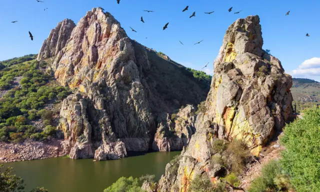 Felsige Berge und ein Fluss in Extremadura, Spanien. Geier kreisen am blauen Himmel über der rauen Landschaft.