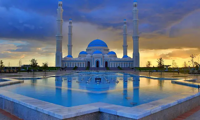 Die Hazrat-Sultan-Moschee in Astana, ein architektonisches Meisterwerk, spiegelt sich im Wasser eines Brunnens unter einem dramatischen Abendhimmel.