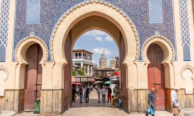 Prachtvolles Tor in Fes Stadt mit blau-goldenen Mustern. Menschen beleben den Platz dahinter.
