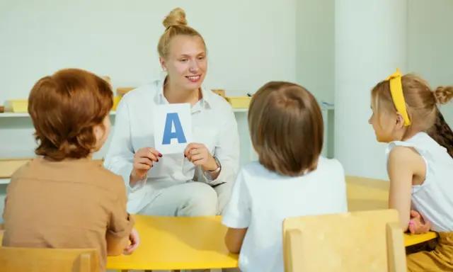 Nauczycielka pokazuje literę "A" grupie dzieci. Jedno z dzieci, 6-latek, uważnie słucha.