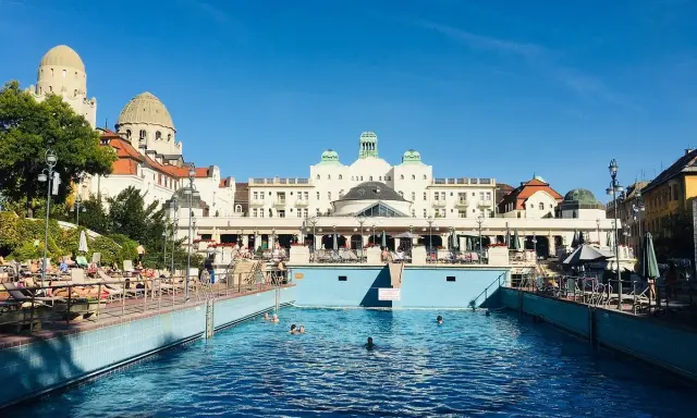Soligt utomhuspool vid ett termalbad i Budapest. Människor solar och badar i det blå vattnet framför en imponerande byggnad.