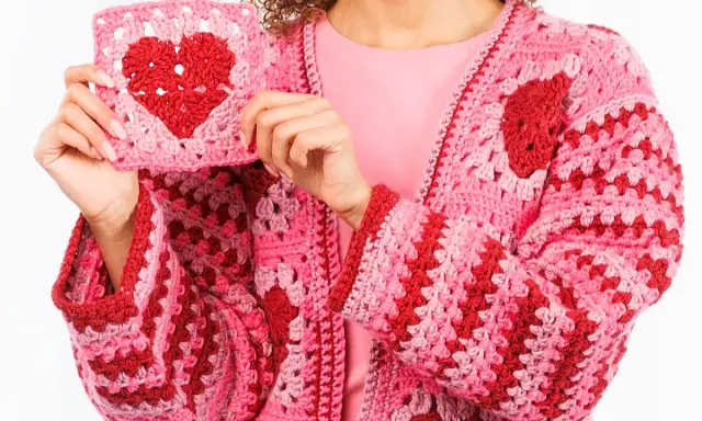 Una mujer muestra un cardigan tejido a mano, rosa y rojo, con un corazón de ganchillo. Este es un ejemplo de qué es un cardigan.