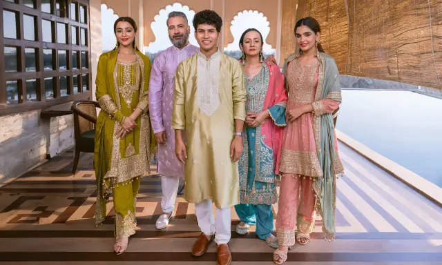 A family poses in vibrant traditional clothing from India. The women wear ornate salwar kameez sets in olive green, teal, and pink, while the men are dressed in elegant kurtas.