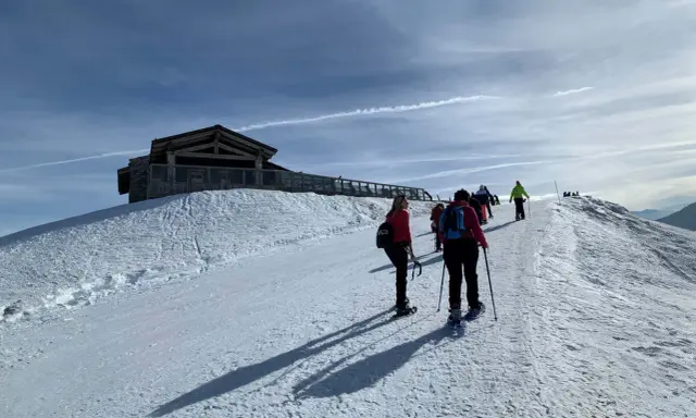 Gruppo di escursionisti con ciaspole su un sentiero innevato, con un rifugio in legno in cima a una collina. Posti belli in montagna Piemonte inverno.