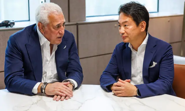 Koji Watanabe conversa con un hombre mayor, ambos con trajes azules. Un modelo de coche deportivo negro se ve en el fondo.