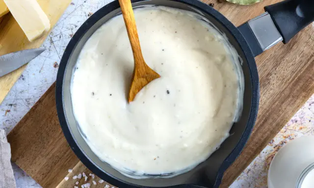 Cremige Bechamelsauce in einer Pfanne, bereit für die Lasagne. Ein Holzlöffel rührt die sämige Masse.
