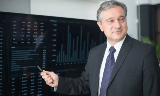Mężczyzna w garniturze wskazuje na ekran z danymi finansowymi, wyjaśniając, kto płaci doradcy kredytowemu.