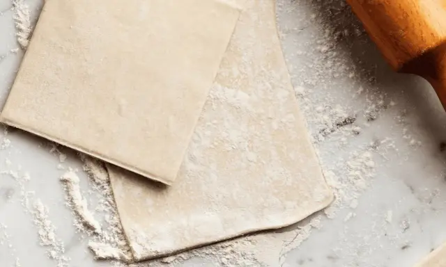 Pâte feuilletée prête à être travaillée, saupoudrée de farine sur un plan de travail en marbre. Un rouleau à pâtisserie en bois est visible sur la droite.