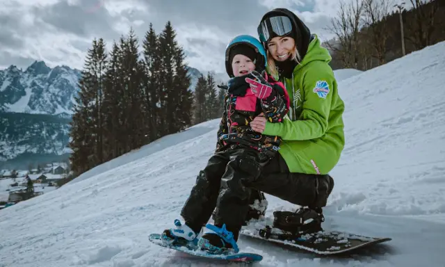 Uśmiechnięta instruktorka na stoku narciarskim dla dzieci trzyma małego snowboardzistę.