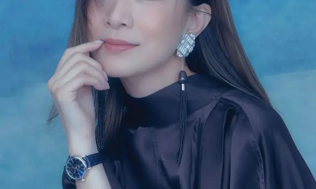 A woman with a thoughtful expression, her finger near her lips, wears a dark satin dress, a striking earring, and a blue-faced watch. She looks xa thi man.