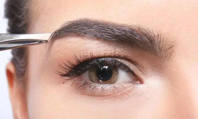 Une pince à épiler retire délicatement un poil du sourcil, affinant la ligne pour un regard parfait.