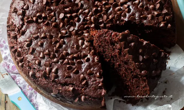 Torta al cioccolato senza uova, soffice e golosa, ricoperta di gocce di cioccolato. Una fetta mostra la sua morbidezza.