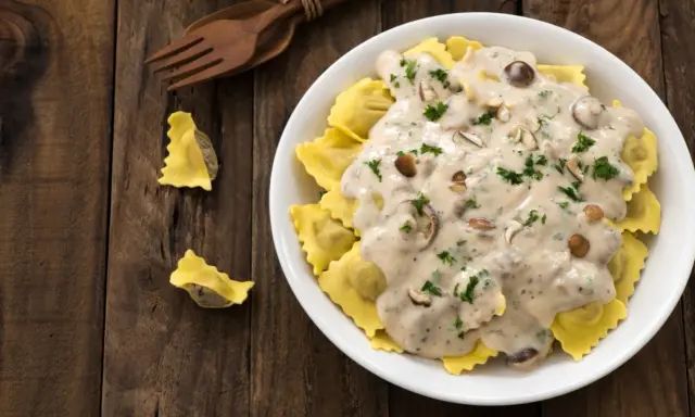 Ravioli w kremowym sosie śmietanowym z grzybami i natką pietruszki, podane na drewnianym stole.