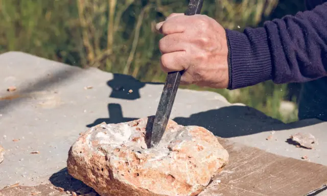 Dłoń z dłutem pracuje nad kamieniem, tworząc pył. To jak wiercenie w granicie z udarem czy bez – wymaga precyzji i siły.