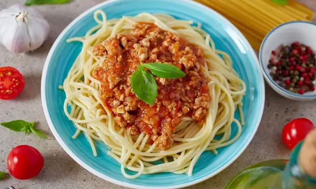 Pyszne spaghetti z bogatym sosem bolognese, udekorowane listkiem bazylii. Obok czosnek, pomidorki i przyprawy.