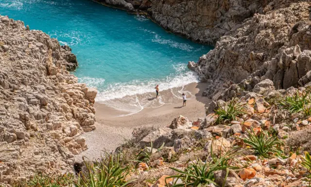 Malownicza zatoka z turkusową wodą i piaszczystą plażą, otoczona skalistymi klifami. Dwie osoby spacerują po brzegu. Chania plaże zachwycają.