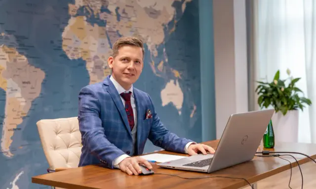 Mężczyzna w garniturze przy biurku z laptopem, na tle mapy świata. Tak wyglądają studia doktoranckie w dzisiejszych czasach – globalne spojrzenie i praca z technologią.