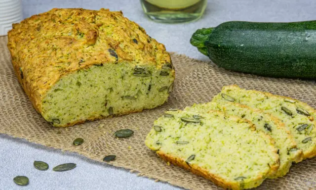 Puszysty chleb z cukinii, z pestkami dyni, obok świeżej cukinii i butelki oleju. Idealny na zdrowe śniadanie.