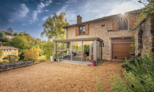 Maison en pierre avec véranda moderne, illustrant la différence entre véranda et extension. Le soleil brille sur le paysage verdoyant.