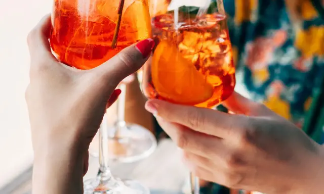Zwei Hände halten Aperol Spritz Gläser mit Eis und Orangenscheiben. Ein perfektes Aperol Rezept für den Sommer.