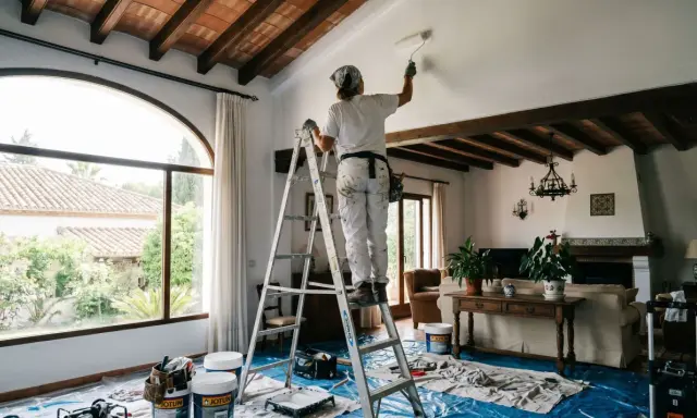 Persona en escalera aplica pintura blanca en techo, parte de un proyecto de alisado de paredes.