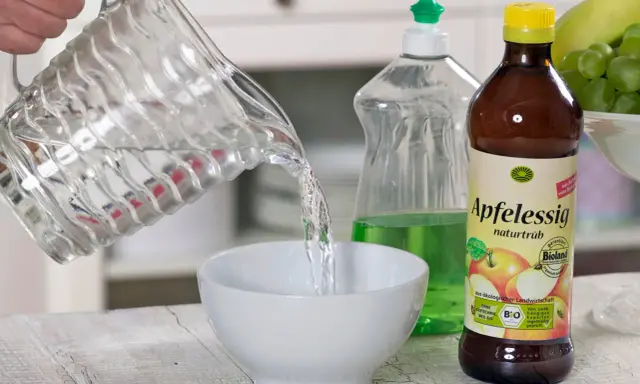 Wasser wird in eine Schüssel gegossen, um eine Fruchtfliegenfalle zu bauen. Apfelessig und Spülmittel sind bereit.