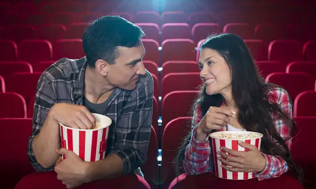 Para w kinie, zajadając popcorn, patrzy na siebie z uśmiechem. Ich spojrzenia sugerują, że to nie tylko film ich interesuje, ale też wzajemna bliskość.