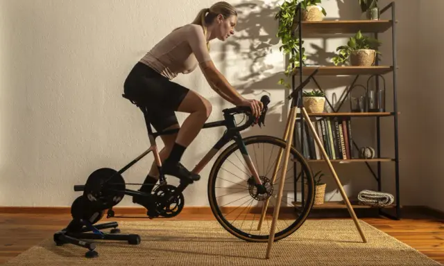 Frau trainiert auf einem Heimtrainer, was ist spinning? Sie tritt kräftig in die Pedale, während im Hintergrund ein Regal mit Pflanzen und Büchern steht.