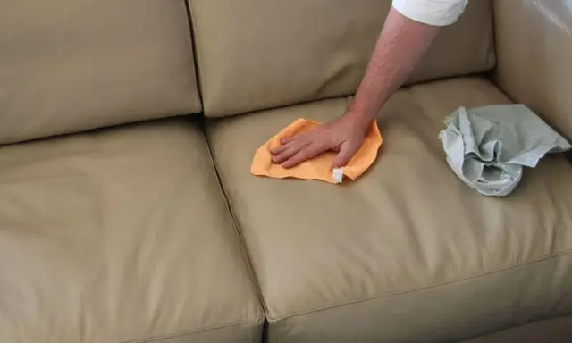 Mano che pulisce un divano in ecopelle beige con un panno arancione. Un altro panno grigio è appoggiato sul divano.