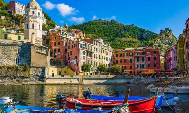 Colorate case a picco sul mare, barche rosse e blu in primo piano, un campanile svetta sulla collina. Un sogno di vacanze 5 Terre.