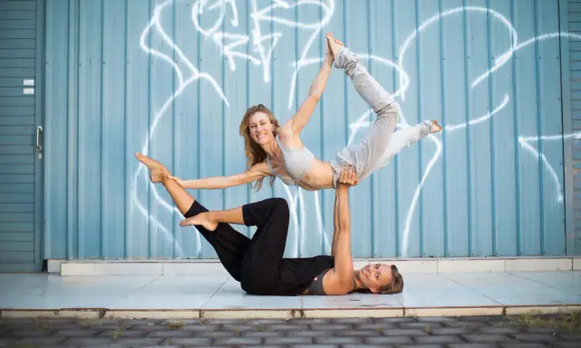 Två kvinnor utför acro yoga framför en blå vägg med graffiti. Den ena ligger på rygg och lyfter den andra som är i en böjd position.