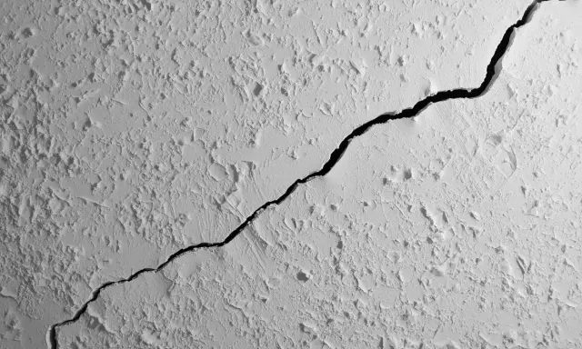 Gros plan sur une fissure traversant un plafond texturé. La fissure est sombre et profonde, contrastant avec la surface blanche et irrégulière.