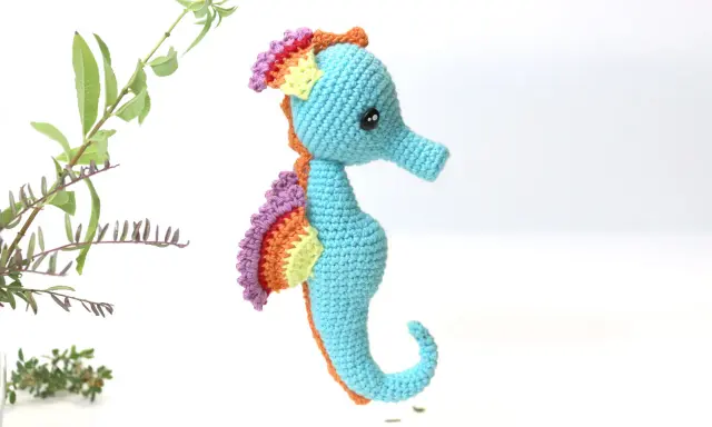 Caballito de mar amigurumi patron gratis. Un adorable caballito de mar tejido a crochet, con aletas arcoíris y cuerpo azul, listo para navegar en tu imaginación.