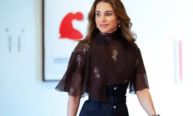 Queen Rania fashion: A woman in a dark brown, floral-patterned top with a high neckline and flared sleeves, paired with a dark skirt.