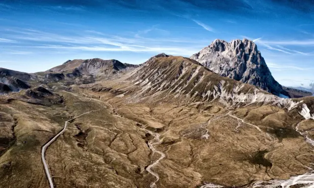 Vasto panorama del **campo imperatore gran sasso** con strade tortuose che si snodano tra montagne brulle e un cielo azzurro intenso.