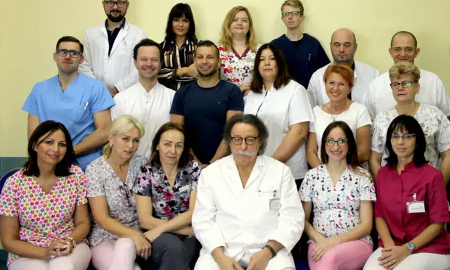 Grupa lekarzy i pielęgniarek w białych fartuchach i kolorowych strojach medycznych, pozująca do zdjęcia.