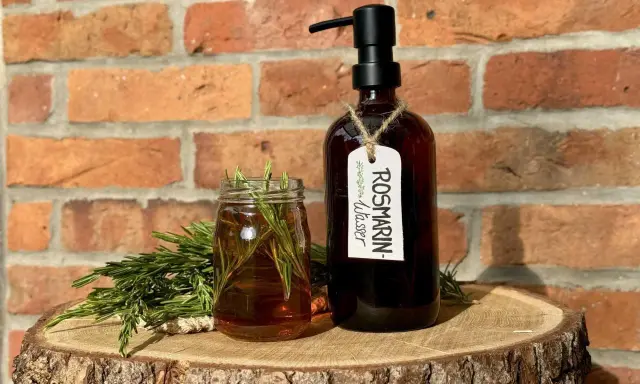 Zutaten für Rosmarinwasser Haare selber machen: frischer Rosmarin, ein Glas mit Flüssigkeit und eine dunkle Flasche mit Pumpspender.