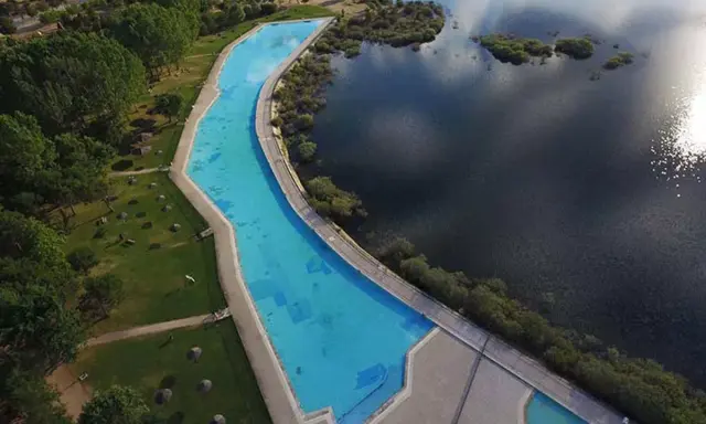 Vista aérea de la piscina natural de Riosequillo, con su agua azul brillante bordeada por césped verde y un lago oscuro.