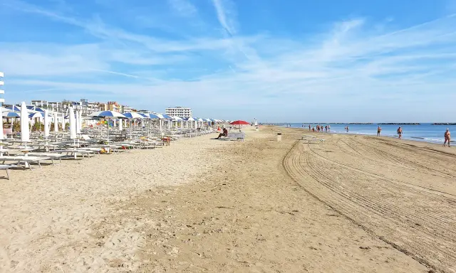 Spiaggia di Ravenna Lido Adriano con file di ombrelloni e lettini, bagnanti che passeggiano sul bagnasciuga sotto un cielo azzurro.