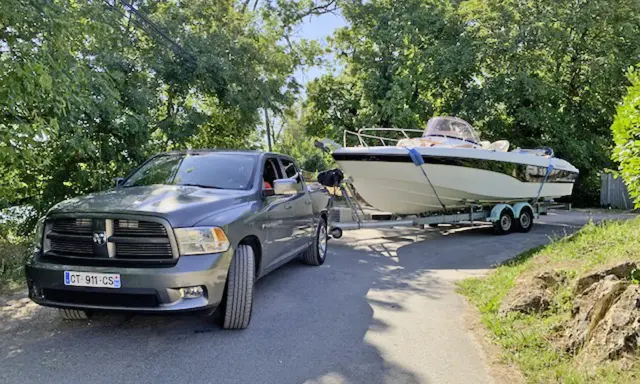 Un pick-up gris est prêt à tracter un bateau de 7m sur sa remorque, sous un ciel bleu et entouré de verdure.
