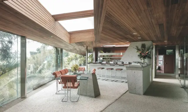 Interno della Sheats Goldstein Residence con soffitto in legno, ampie vetrate, zona pranzo con tavolo in vetro e sedie in pelle arancione, e un bancone bar in cemento con sgabelli.